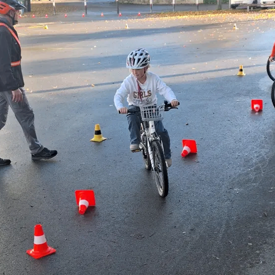 Ein junger Junge mit Helm fährt auf einer nassen Straße ein Fahrrad und navigiert um Verkehrskegel herum. Ein Erwachsener geht neben ihm. Im Hintergrund sind Autos geparkt.