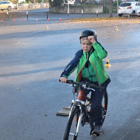 Ein junger Junge mit Helm fährt auf einer nassen Straße mit einem Fahrrad, umgeben von Verkehrskegeln. Hinter ihm sind Autos in der Nähe eines Gebäudes mit Bäumen geparkt.