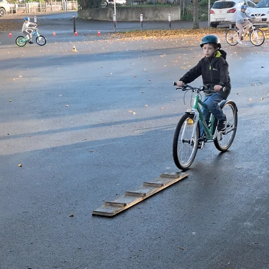 Ein junger Junge fährt mit seinem Fahrrad über eine Holzrampe auf einer asphaltierten Straße, während zwei andere Kinder im Hintergrund mit ihren Fahrrädern fahren.
