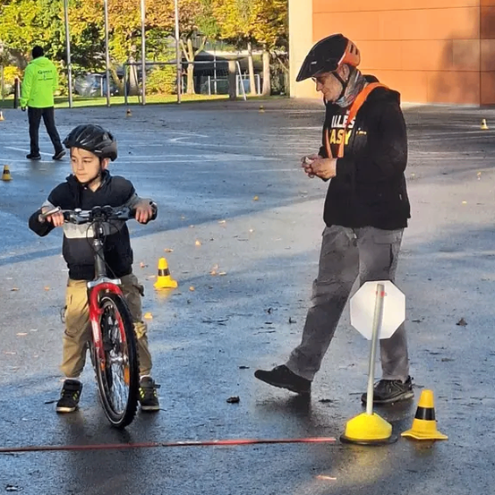 Ein junger Junge fährt ein Fahrrad, begleitet von einem Erwachsenen. Sie befinden sich in einem Außenbereich mit nasser Fahrbahn, umgeben von Verkehrskegeln. Bäume und Gebäude sind im Hintergrund zu sehen.