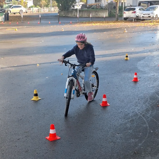 Ein Mädchen fährt mit einem Fahrrad durch eine nasse Straße und navigiert um Verkehrskegel herum, während es einen Helm trägt. Hinter ihr sind mehrere Autos geparkt, und Bäume säumen die Straße.