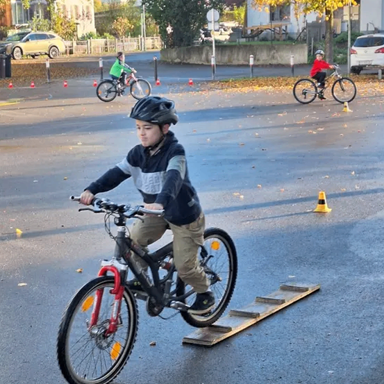 Ein Junge fährt mit seinem Fahrrad über eine Holzrampe in einem Parkplatz mit Verkehrskegeln. Hinter ihm fahren zwei weitere Kinder auf Fahrrädern. Bäume und Häuser sind im Hintergrund.