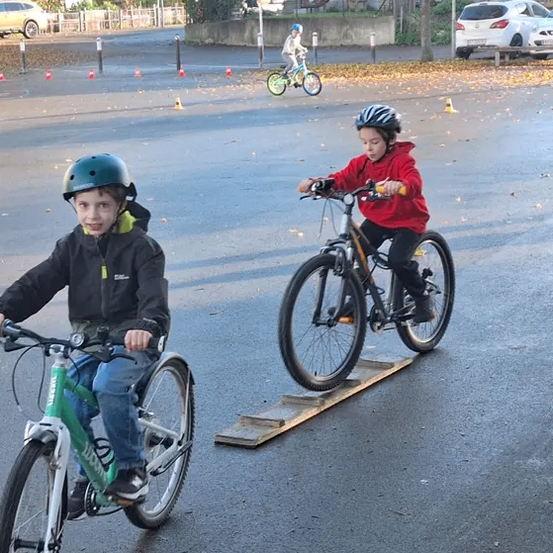 Kinder fahren auf einem gepflasterten Weg mit Fahrrädern und tragen Helme. Ein Kind fährt auf einer hölzernen Rampe. Verkehrskegel markieren das Gebiet. Im Hintergrund fahren andere Kinder mit Fahrrädern auf der Straße. Autos sind am Straßenrand geparkt.
