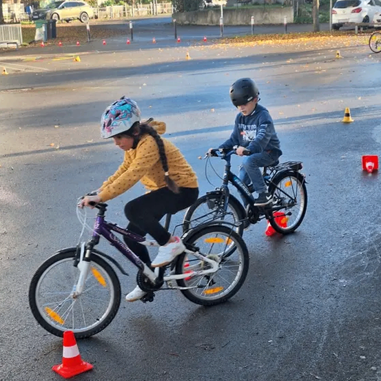 Zwei Kinder fahren auf einer nassen Straße mit Fahrrädern, umgeben von Verkehrskegeln. Ein Kind trägt einen Helm, während das andere einen Rucksack trägt. Im Hintergrund befinden sich Gebäude und Autos.