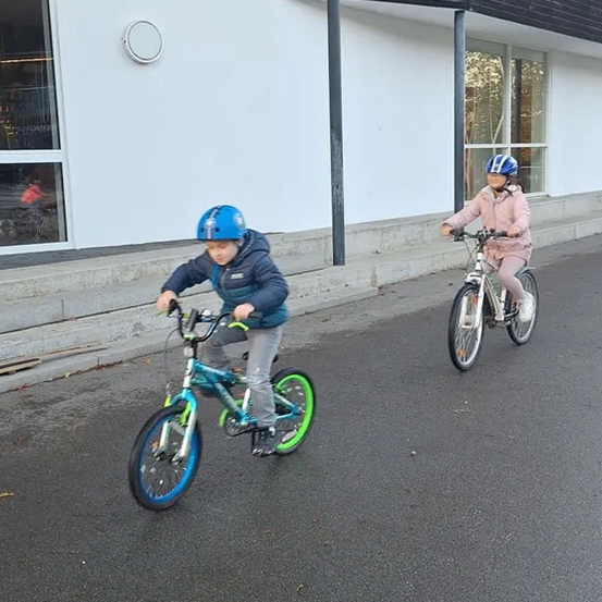 Zwei Kinder fahren auf einer Straße vor einem Gebäude mit Glasfenstern mit ihren Fahrrädern.