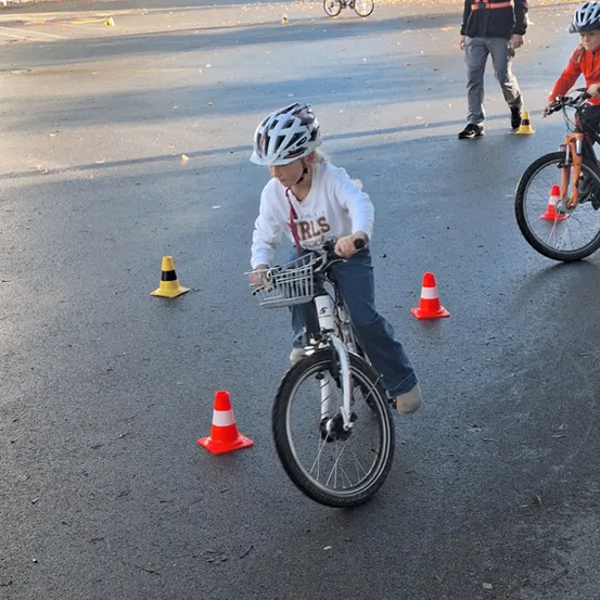 Ein Kind mit Helm fährt ein Fahrrad auf einem Parkplatz mit Verkehrskegeln und einem Mann in Sicherheitsausrüstung in der Nähe. Andere Kinder und Fahrräder sind im Hintergrund zu sehen.