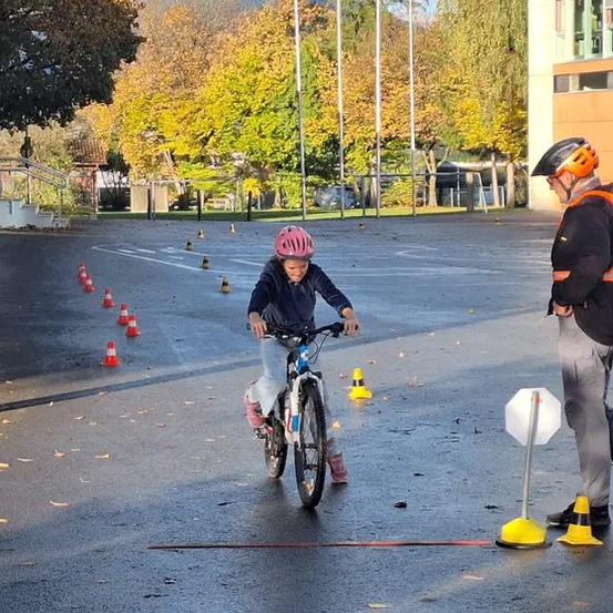 Ein Kind fährt mit einem Fahrrad durch einen Parcours, der mit Hütchen markiert ist. Das Kind trägt einen Helm und Turnschuhe. Ein Erwachsener steht in der Nähe und trägt eine Warnweste und einen Helm. Bäume und ein Gebäude sind im Hintergrund zu sehen.
