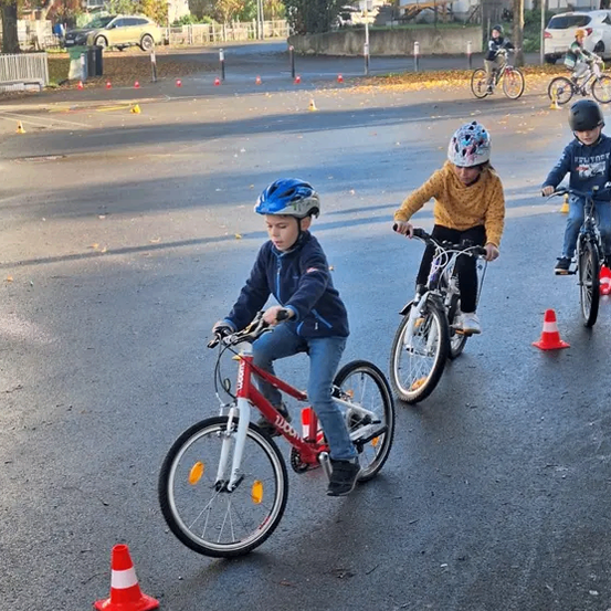 Drei Kinder fahren auf einer Straße mit Verkehrskegeln auf Fahrrädern. Ein Kind trägt einen blauen Helm. Im Hintergrund befinden sich Gebäude und Bäume.