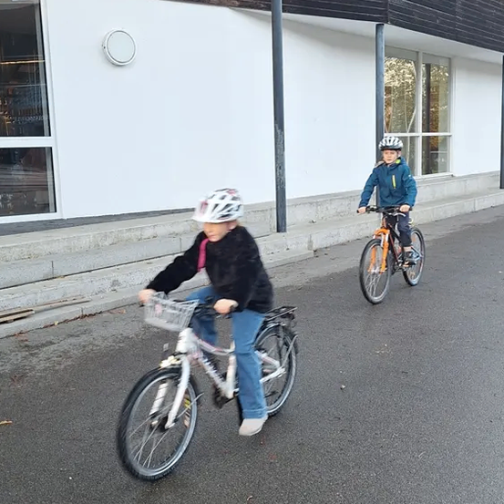 Zwei Kinder fahren vor einem Gebäude mit Glasfenstern und einer grauen Wand auf Fahrrädern.