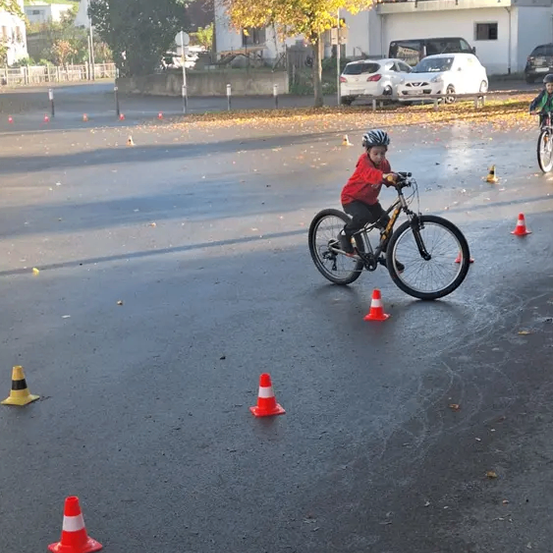 Ein Kind fährt auf einer nassen Straße mit Verkehrskegel darum ein Fahrrad, vor einem Gebäude mit geparkten Autos.