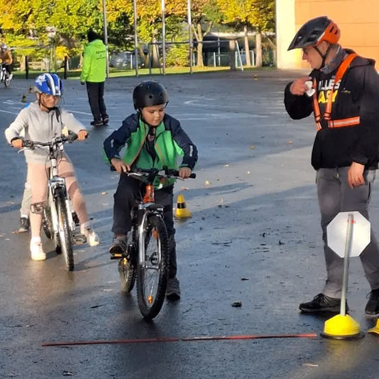Drei Kinder fahren auf einer nassen Straße mit Fahrrädern. Eines trägt eine grüne Jacke, ein anderes einen blauen Helm. Ein Erwachsener steht mit einem Schild in der Nähe. Bäume und Gebäude sind im Hintergrund.