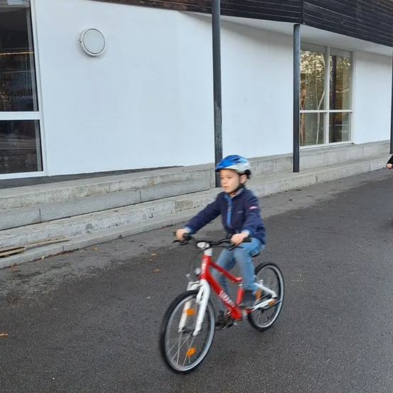 Ein Kind fährt einen roten Fahrrad die Straße hinunter und trägt einen Helm. Hinter ihm steht ein Gebäude mit Glasfenstern und Stufen.