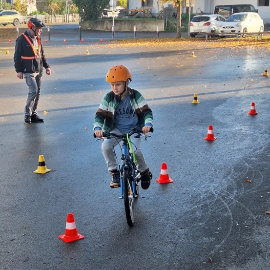 Ein Kind in einem Helm fährt ein Fahrrad durch einen nassen Parcours, der von orangefarbenen Kegeln markiert ist. Ein Mann steht in der Nähe. Die Szene umfasst Autos, die in einem Parkplatz abgestellt sind.