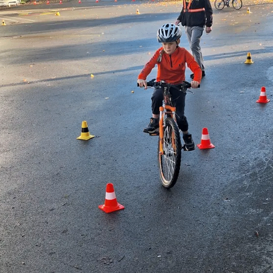 Ein junges Kind fährt mit einem Fahrrad durch einen Parcours, der mit orangefarbenen und weißen Kegel markiert ist, auf einer nassen Straße. Ein Erwachsener folgt ihm dicht dahinter.