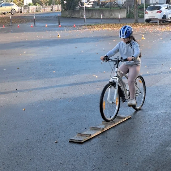 Ein Mädchen mit Helm fährt ein Fahrrad über eine Holzplanke auf einer nassen Straße mit Verkehrskegeln und geparkten Autos im Hintergrund.