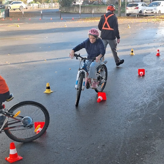 Ein Kind in einem Helm fährt ein Fahrrad durch einen Parcours mit Verkehrskegel auf der Straße. Ein Mann in einer reflektierenden Weste geht neben ihnen.