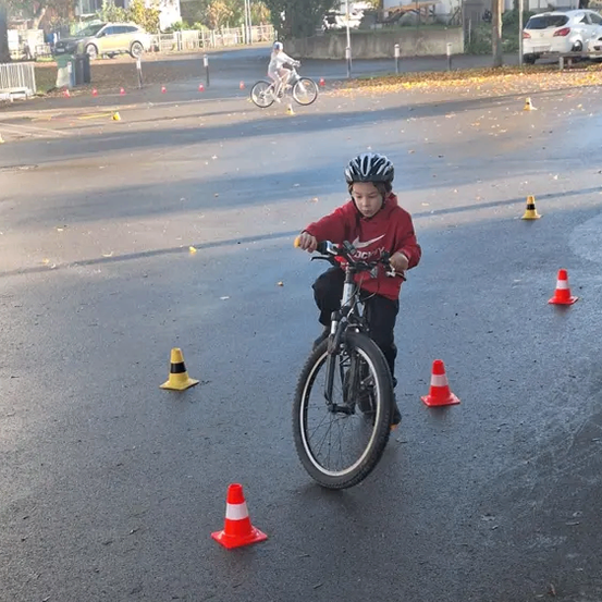 Ein junges Kind fährt auf einer Straße mit Verkehrskegeln ein Fahrrad. Ein weiterer Radfahrer fährt im Hintergrund.