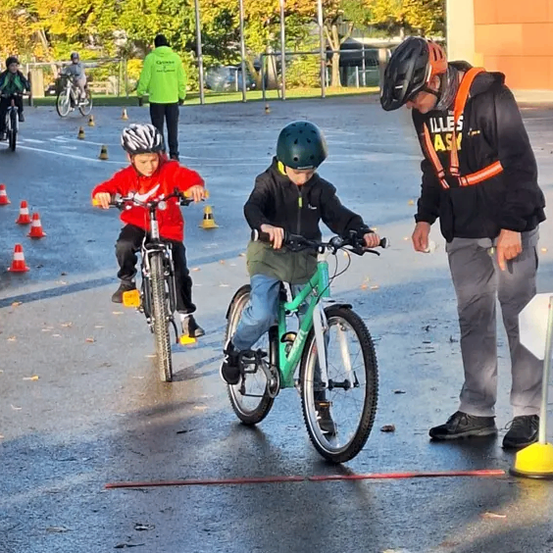 Zwei Kinder fahren auf einem Freigelände mit Fahrrädern, während ein Mann sie anleitet. Mehrere Verkehrskegel sind auf der Straße platziert.