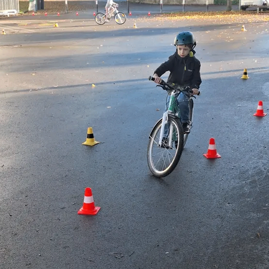 Ein Kind fährt auf einem Fahrrad in einem Parkplatz, umgeben von Verkehrskegel. Ein weiteres Kind fährt in der Ferne. Bäume und geparkte Autos sind im Hintergrund zu sehen.