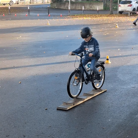 Ein junger Junge fährt mit einem Helm auf einem gepflasterten Weg mit einem Fahrrad über eine Holzplanke, umgeben von Verkehrskegeln.