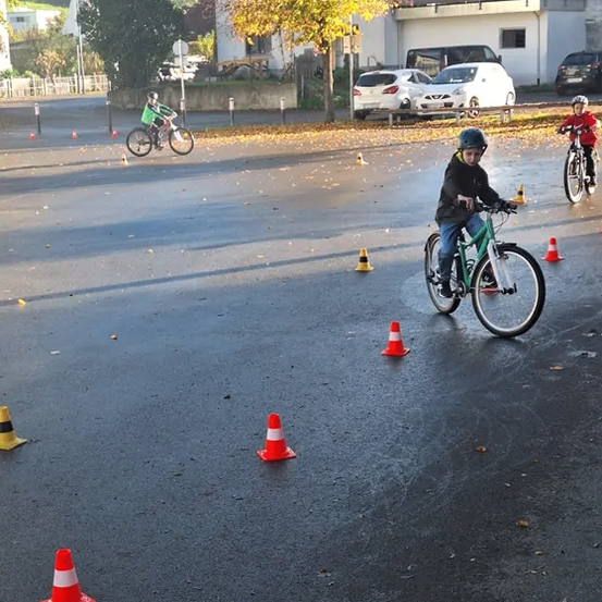 Drei Kinder fahren mit Fahrrädern durch eine nasse Straße und navigieren um orangefarbene und gelbe Kegel herum. Ein Kind trägt ein grünes Shirt, ein anderes einen roten Helm. Autos sind in der Nähe geparkt, und Gebäude sind im Hintergrund zu sehen.