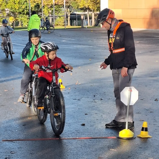Ein Outdoor-Fahrradkurs mit mehreren Kindern, die Helme tragen und ihre Fahrräder fahren. Ein Mann hält ein Stoppschild vor sie.