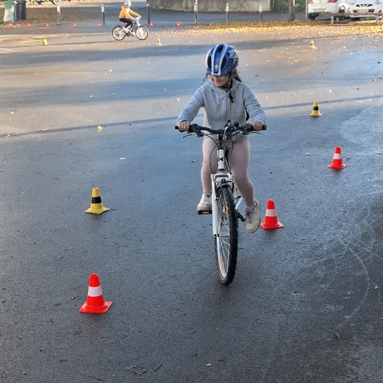 Ein junges Mädchen fährt mit Helm und Turnschuhen auf einer nassen Straße mit dem Fahrrad, umgeben von Verkehrskegel. Ein weiterer Radfahrer ist hinter ihr. Im Hintergrund befinden sich Gebäude, Autos und Bäume.