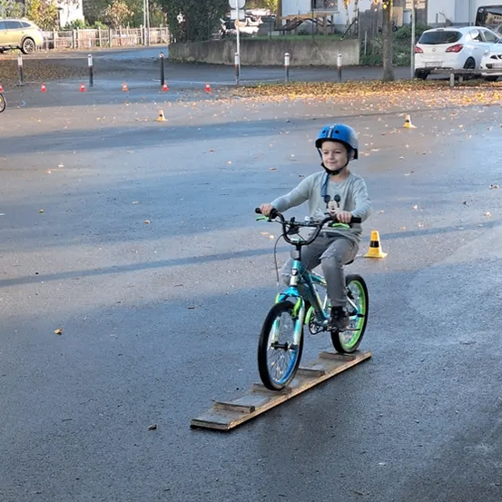 Ein Junge fährt mit einem Fahrrad auf einer hölzernen Rampe in einem Parkplatz. Er trägt einen Helm und Turnschuhe. Mehrere Verkehrskegel sind verstreut.