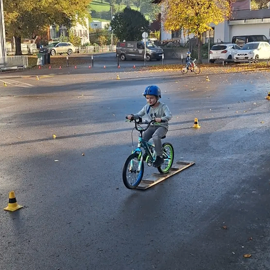 Ein Kind mit Helm fährt auf einer Straße mit Verkehrskegeln auf einem Fahrrad und ein anderes Kind fährt dahinter.