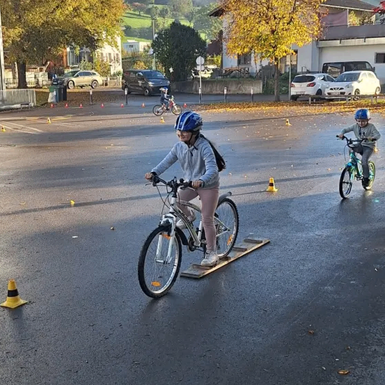 Zwei Kinder fahren auf einer nassen Straße mit Verkehrskegeln auf Fahrrädern. Sie tragen Helme und fahren in der Nähe eines Gebäudes mit geparkten Autos.