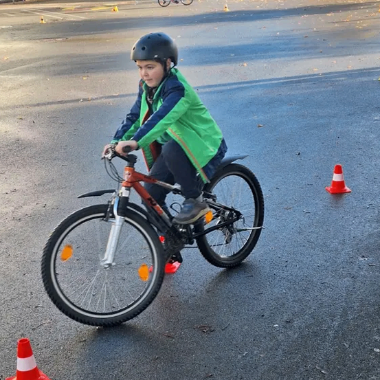 Ein Kind fährt mit einem Helm auf einem Fahrrad zwischen Verkehrskegel auf einer asphaltierten Straße. In der Ferne fährt ein weiteres Kind ein Fahrrad. Bäume und ein Auto sind im Hintergrund zu sehen.