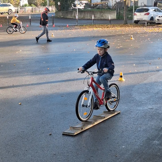 Ein Junge fährt auf einem Holzbrett auf der Straße mit dem Fahrrad. Hinter ihm geht eine Person. Einige Verkehrskegel sind auf der Straße platziert. Ein Auto ist auf der rechten Seite geparkt.