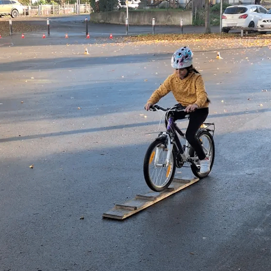 Ein Mädchen fährt mit einem Fahrrad auf einer Rampe in einer leeren Straße, trägt einen Helm und eine gelbe Jacke. Es gibt orangefarbene Verkehrskegel und geparkte Autos im Hintergrund.