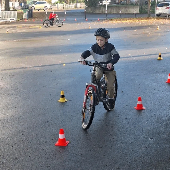 Ein Kind fährt zwischen orangefarbenen Verkehrskegeln auf einem offenen Gelände ein Fahrrad. Ein weiteres Kind fährt im Hintergrund ein Fahrrad. Autos sind am Straßenrand geparkt.