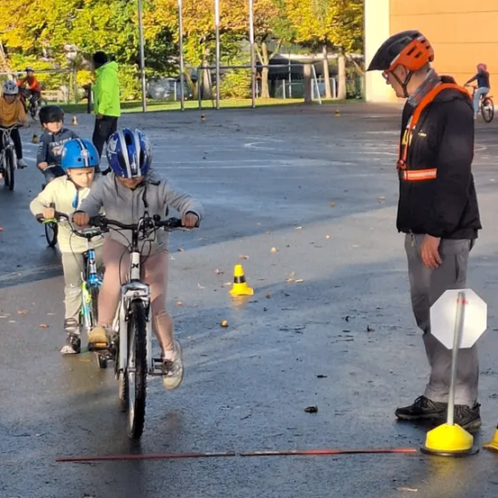 Kinder fahren auf einer nassen Oberfläche mit Fahrrädern, wobei ein Mann beaufsichtigt. Helme sind getragen, und Verkehrskegel sind platziert. Bäume mit gelben Blättern und ein Gebäude sind im Hintergrund.