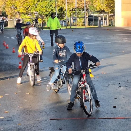 Eine Gruppe von Kindern, die auf einer nassen Straße mit Verkehrskegeln und einem vorbeigehenden Mann Fahrrad fahren.