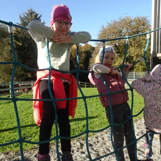 Drei Kinder klettern auf einem Netz im Freien. Das Mädchen in der Mitte trägt eine rosa Kopfband und eine Brille. Die anderen beiden tragen Jacken.