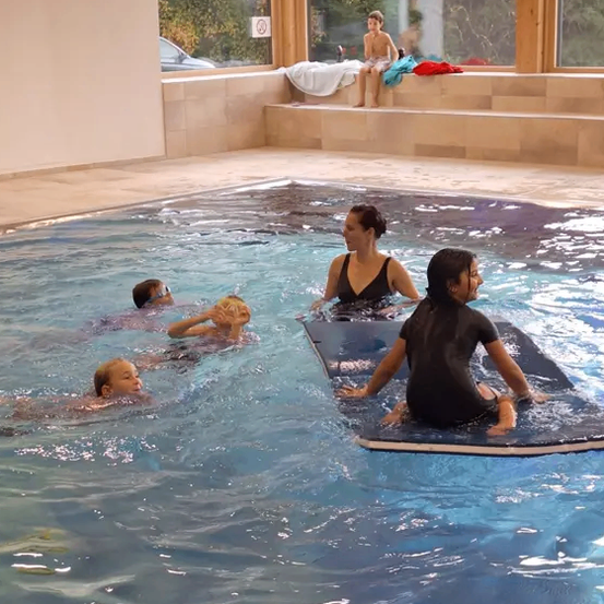 Eine Gruppe von Kindern und Erwachsenen schwimmt in einem Pool, wobei zwei Frauen ihnen helfen. Ein Kind sitzt auf einem Surfbrett.