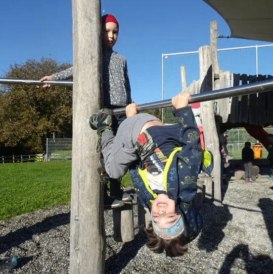 Zwei Kinder spielen auf einem Spielplatz, eines hängt kopfüber an einer Stange, das andere steht auf einem Pfosten. Im Hintergrund sind ein Rasenbereich und Bäume zu sehen.