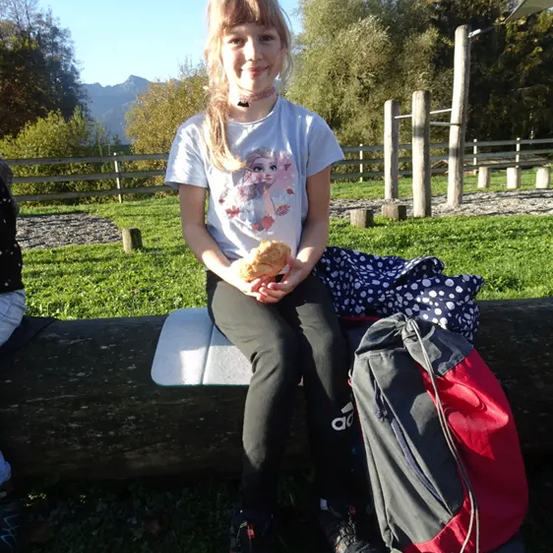 Ein junges Mädchen in einem weißen T-Shirt sitzt auf einem Baumstamm in einem Park, hält ein Sandwich und lächelt. Hinter ihr liegen ein Rucksack und eine Decke auf dem Boden.