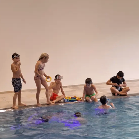 Eine Gruppe von Kindern steht am Rande eines Schwimmbeckens, einige tragen Schwimmbrillen und Badehosen. Ein Junge sitzt am Rand des Beckens, während ein anderer Junge unter Wasser schwimmt.