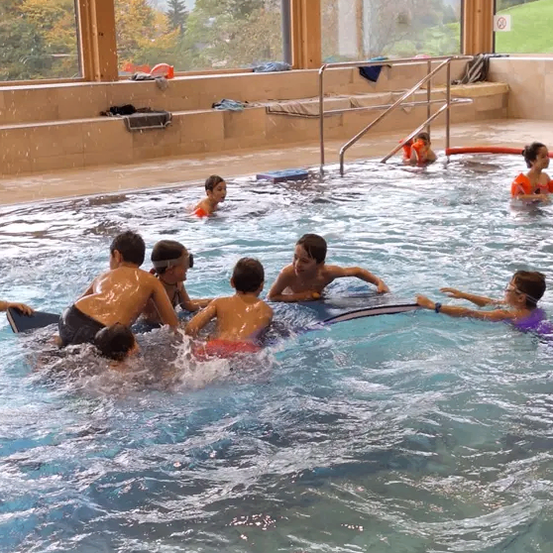 Mehrere Kinder schwimmen in einem Pool mit Blick auf Bäume und Berge vor den Fenstern.