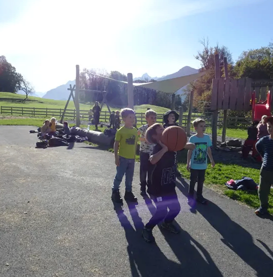 Eine Gruppe von Kindern spielt Basketball auf einem Betonweg. Ein Kind hält einen Basketball. In der Nähe befindet sich ein Spielplatz mit Schaukel und Zaun. Im Hintergrund ein Grasbereich mit Bäumen und Bergen.