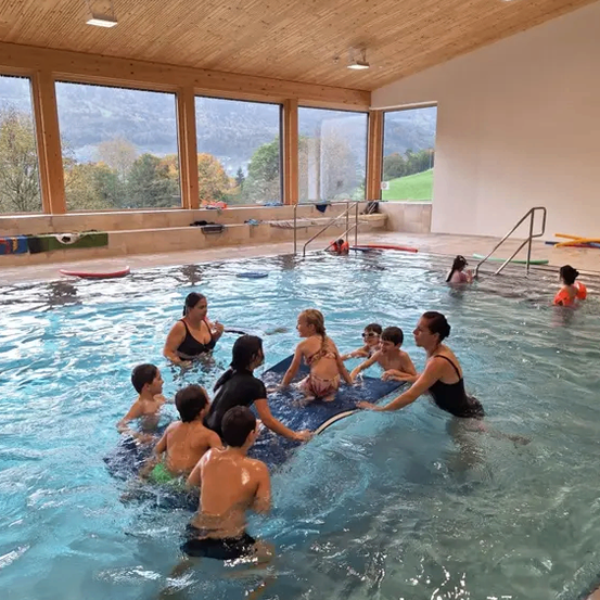 Ein Hallenbad mit Erwachsenen und Kindern beim Schwimmen. Zwei Frauen helfen Kindern auf einem schwimmenden Brett. Fenster und Holzdecke.