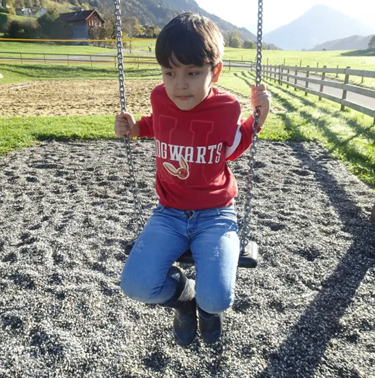 Ein junger Junge in einem roten Shirt und blauen Jeans sitzt auf einer Schaukel in einem Park mit Bergen im Hintergrund.