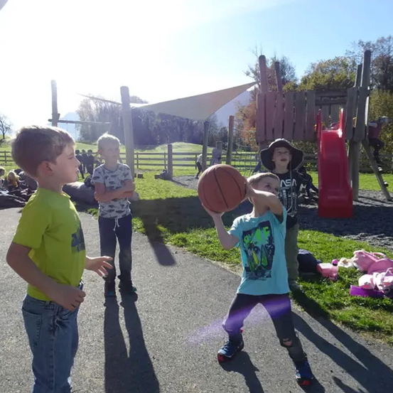 Mehrere Kinder spielen Basketball in einem Park. Ein Junge hält einen Basketball und ist kurz davor zu werfen. Hinter ihm stehen andere Kinder und beobachten.