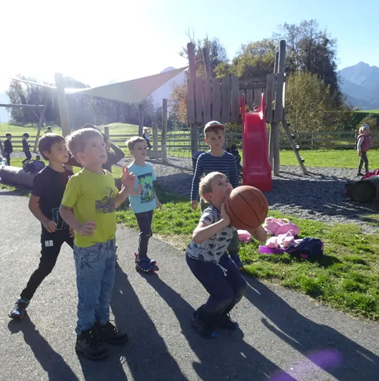 Eine Gruppe von Kindern spielt Basketball in einem Park. Ein Junge hält einen Basketball. In der Nähe gibt es Spielgeräte, eine rote Rutsche und einen Rasenbereich.