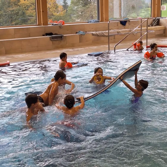 Kinder in orange Schwimmhilfen spielen in einem Pool mit einem großen Surfbrett, zwei halten das Brett, während andere herumspritzen. Glasfenster zeigen einen Blick auf Bäume und Berge.