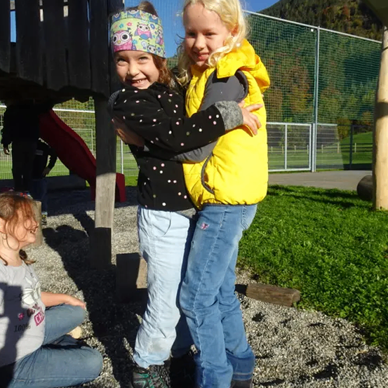 Zwei junge Mädchen umarmen sich an einem sonnigen Tag draußen vor einem Spielplatz mit einer Rutsche und einem Zaun.