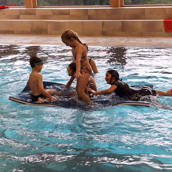 Eine Gruppe von Kindern und Erwachsenen schwimmt in einem Pool mit einer Holzplattform. Die Frau hilft den Kindern. Bäume sind durch die Fenster sichtbar.